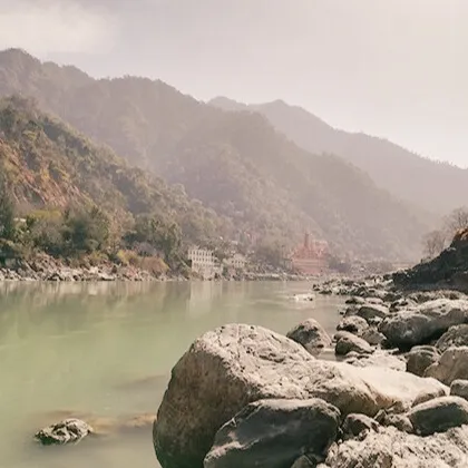Serene view of the Ganges River flowing through the Himalayan foothills near Rishikesh, with rocky banks and misty mountains in the background